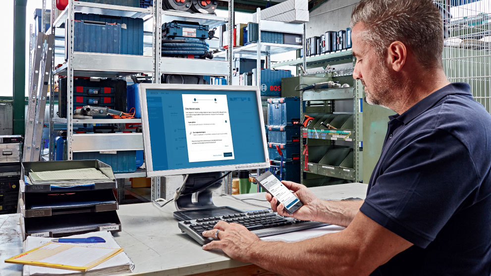 Um homem trabalha em um computador enquanto segura um smartphone na mão. Ao fundo, há prateleiras com caixas de ferramentas e materiais.