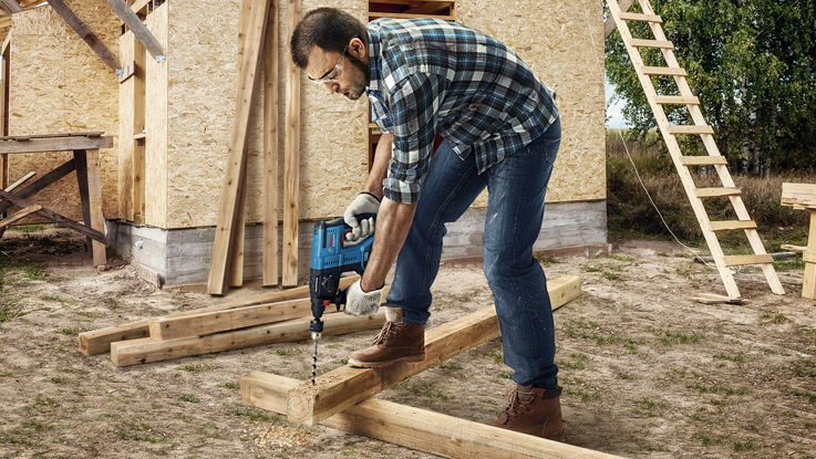 رجل يحمل مثقاب يعمل على ألواح خشبية أمام سقالة.