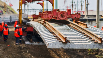 Рабочие в оранжевой одежде укладывают рельсы с помощью крана.