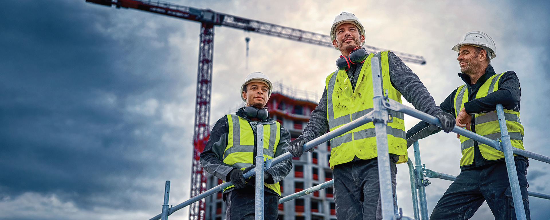Trois ouvriers du bâtiment avec des casques et des gilets de sécurité se tiennent sur un échafaudage.