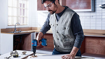 Muž se ochrannými brýlemi vrtá modrým nástrojem Bosch do dřevěné desky.