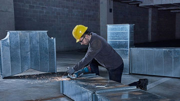 Muž s žlutou helmou řeže kovové části v dílenské oblasti.