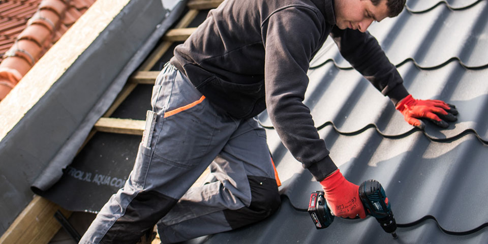Rzemieślnik w odzieży roboczej wkręca na dachu za pomocą wkrętarki akumulatorowej.