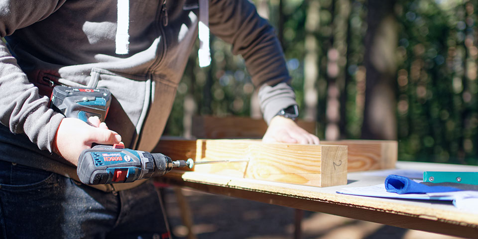 Řemeslník používá akumulátorovou vrtačku Bosch na dřevěném stole v lese.