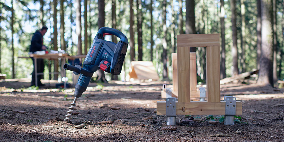 Modrá akumulátorová vrtačka Bosch Professional stojí na lesní půdě vedle dřevěného rámu.