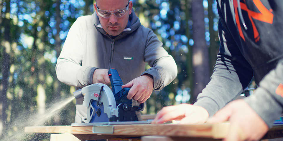 Dva muži pracují s okružní pilou Bosch na dřevěném stole.