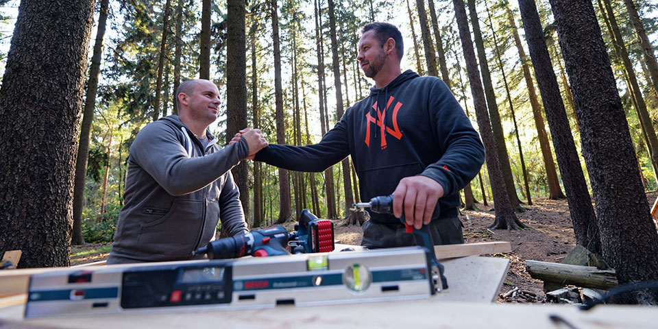 Dva muži si podávají ruce, obklopeni stromy a nástroji.