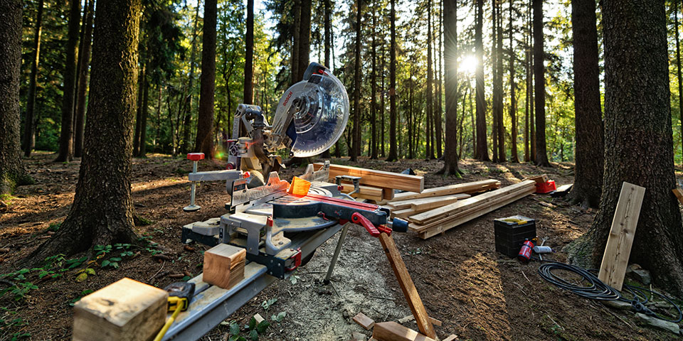 Stolní kotoučová pila stojí v lese, obklopena dřevěnými prkny a stromy.