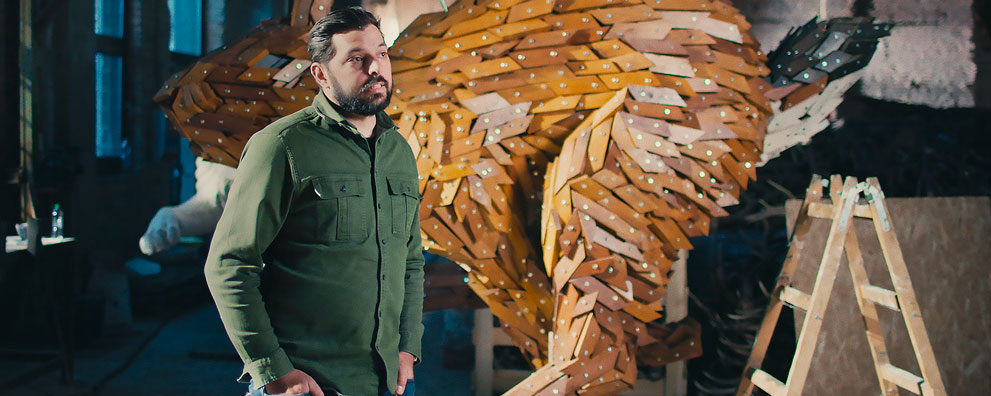 Мужчина в зеленой куртке стоит рядом с деревянной скульптурой животного.