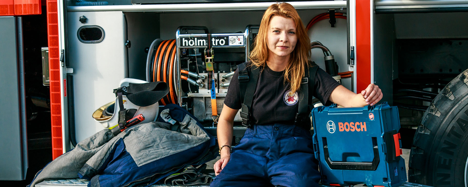 Kobieta w odzieży roboczej siedzi obok niebieskiej skrzynki narzędziowej Bosch.