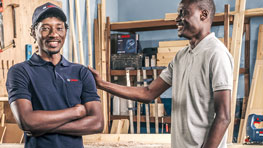 Two men smile in a workshop, surrounded by wood materials.