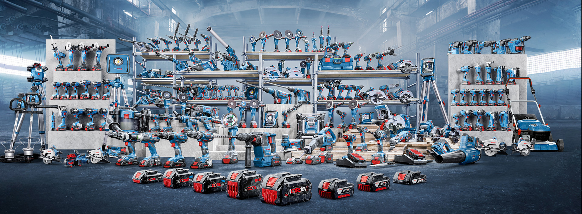 Ett antal blå och röda verktyg och maskiner är prydligt ordnade på hyllor. I förgrunden ligger flera batterier. Verkstaden har en industriell atmosfär med grå väggar och starkt ljus.