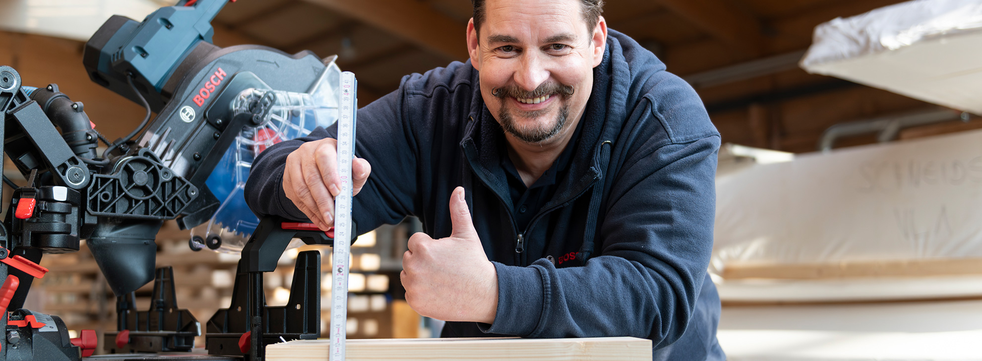 Un artisan sourit à côté d'une scie à onglet Bosch Professional et tient un mètre ruban au-dessus d'un morceau de bois.