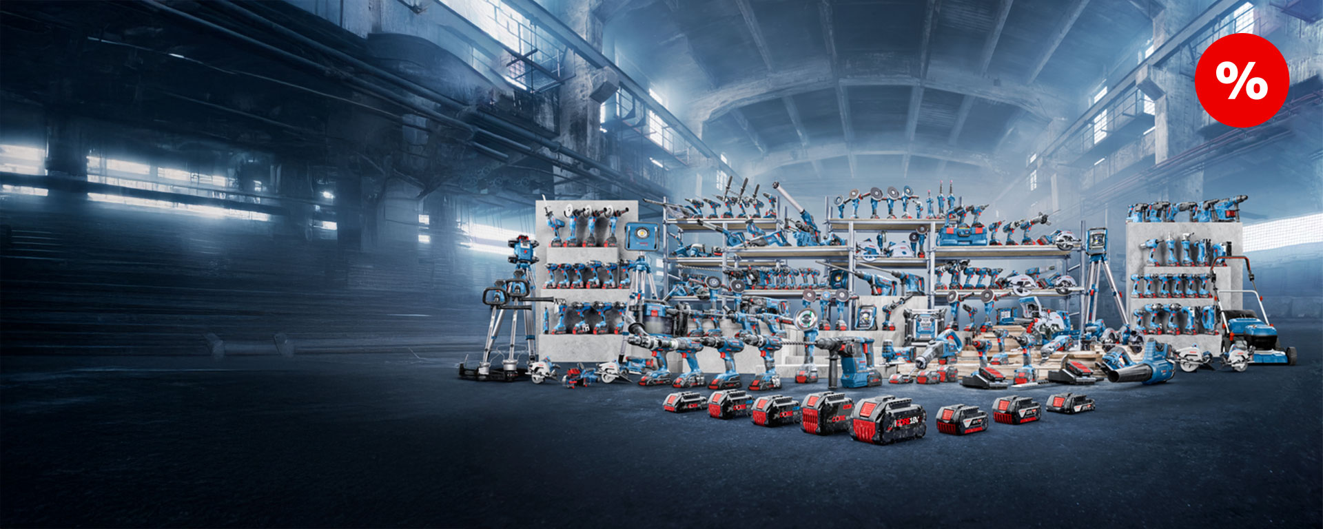 Une large gamme d'outils Bosch Professional bleus est disposée sur des étagères blanches dans une halle industrielle. Certains outils sont éparpillés sur le sol, y compris des appareils sans fil et des accessoires. En arrière-plan, de grandes fenêtres et une atmosphère enfumée sont visibles. Un panneau rouge avec un pourcentage met en avant une remise.