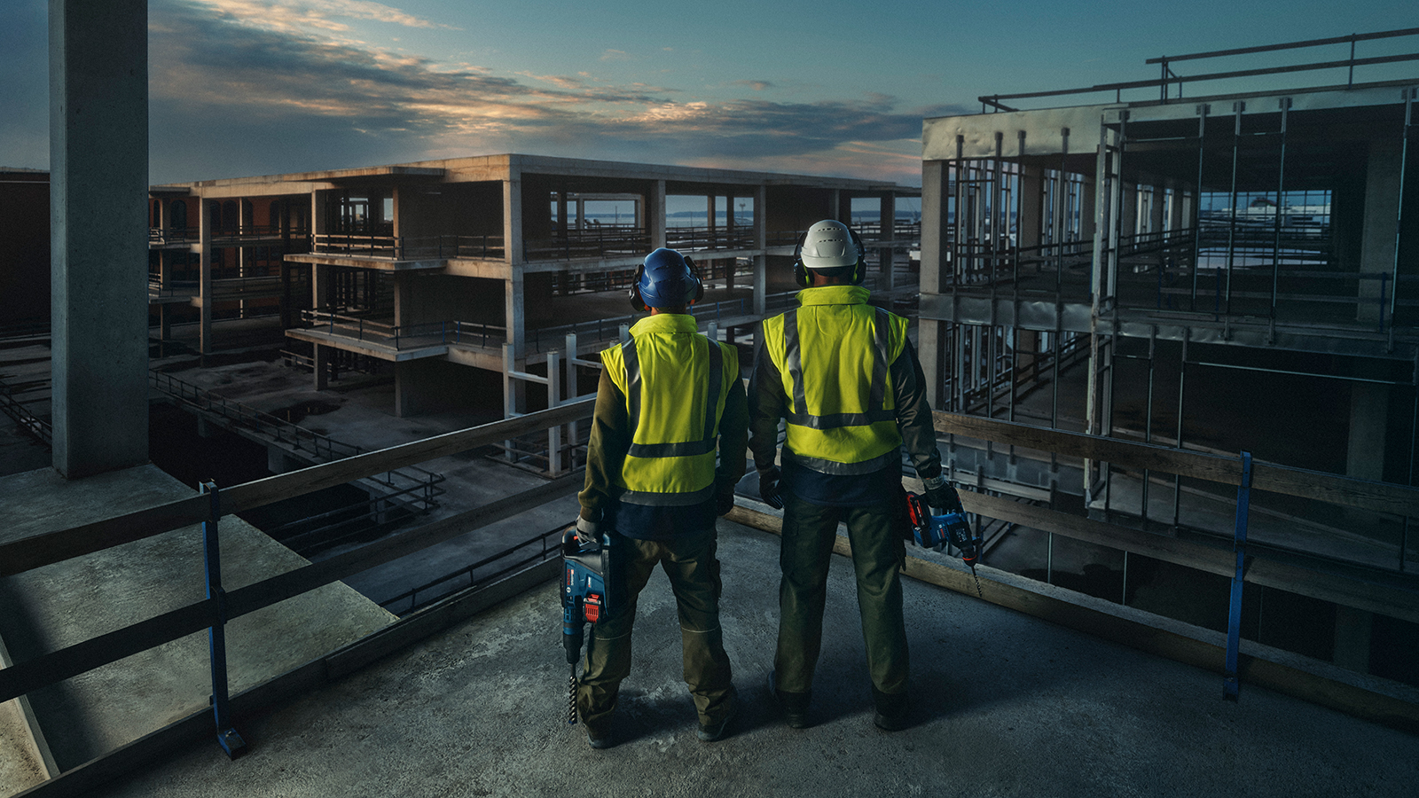 Zwei Bauarbeiter in Warnwesten stehen auf einem Rohbau und betrachten die Baustelle im Abendlicht.