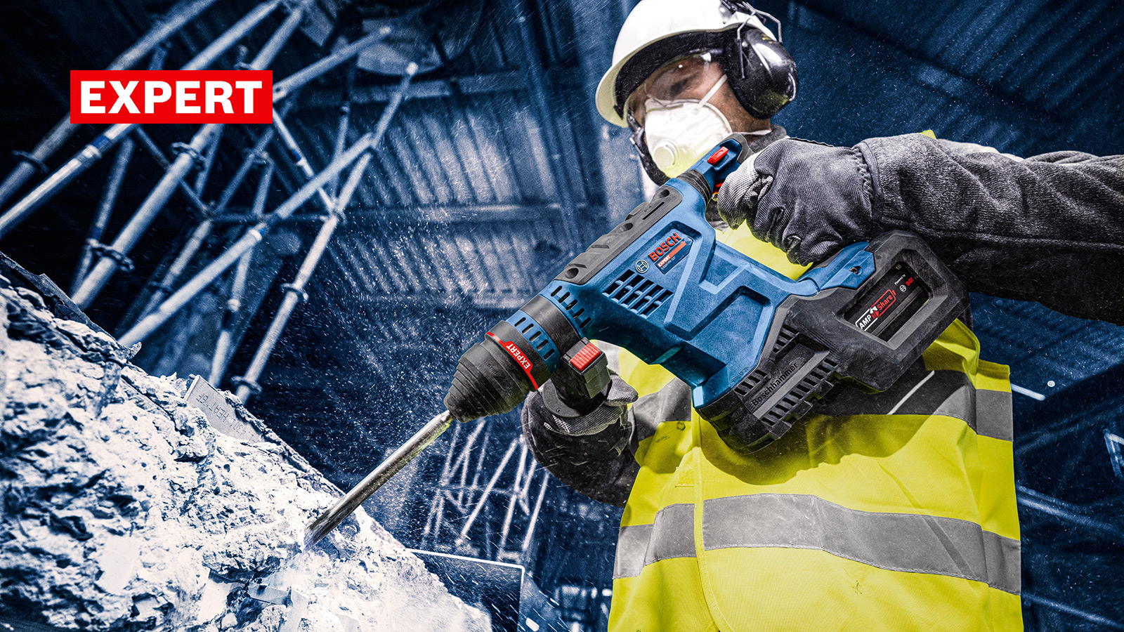 Un trabajador con chaleco de seguridad amarillo y casco utiliza un taladro eléctrico para trabajar el concreto. El polvo vuela.