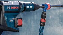 Primer plano de una herramienta Bosch azul con un taladro rojo y negro, sobre una pared gris.