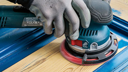 Una mano sostiene una lijadora eléctrica sobre madera, rodeada de mesas de trabajo azules.