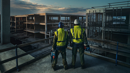 Zwei Bauarbeiter in gelben Sicherheitswesten stehen auf einem Gerüst und blicken auf eine Baustelle bei Sonnenuntergang.