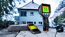 Un dispositivo de termografía y un teléfono inteligente están sobre un ladrillo, al fondo una casa con techo.