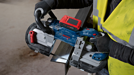 En person i en gul sikkerhedsvest holder en blå båndsav. Baggrunden er uskarp.