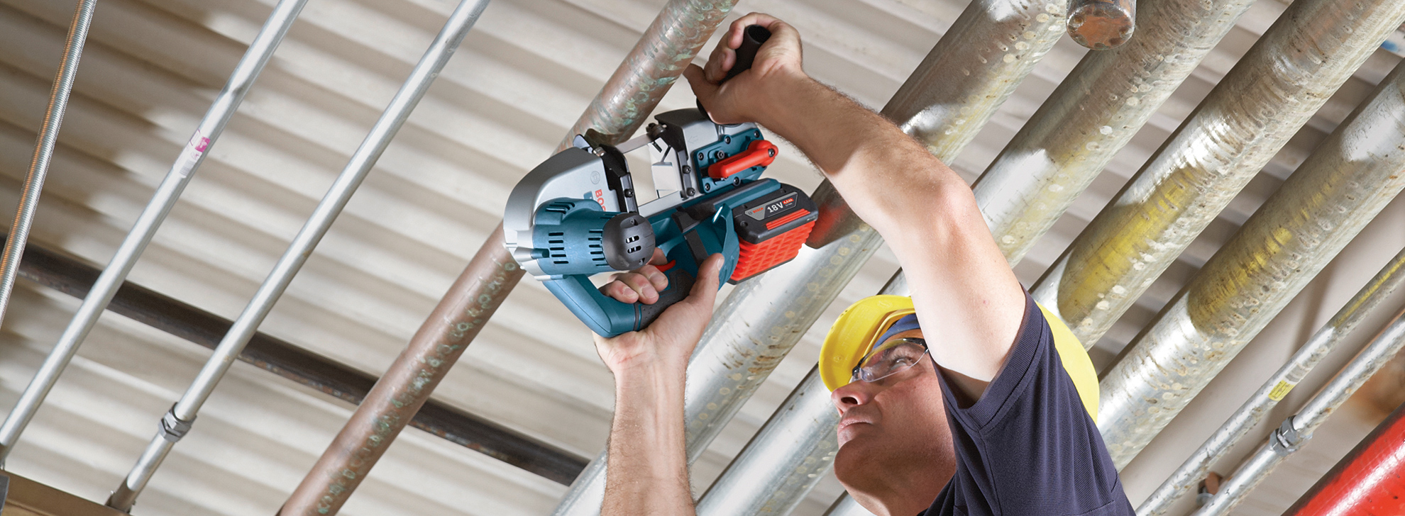 En håndværker bruger en elektrisk sav til at skære metalrør i loftet. Han bærer en gul hjelm og beskyttelsesbriller. Baggrunden viser et struktureret loft og flere metalrør i forskellige farver og størrelser.