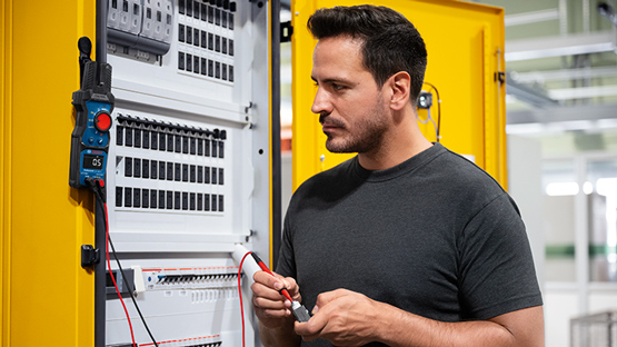 Un homme en t-shirt gris se tient devant un tableau électrique jaune ouvert et vérifie les connexions électriques avec un multimètre. Le tableau est équipé de plusieurs interrupteurs et câbles. L'environnement semble moderne et bien éclairé.