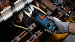 Un multimètre bleu Bosch Professional est utilisé dans un tableau électrique pour tester des câbles. La main porte un gant de travail gris. L'environnement est sombre, avec des câbles visibles de différentes couleurs.