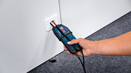 Un testeur de tension Bosch Professional de couleur bleue est tenu contre une prise murale. L'arrière-plan est gris et le sol est recouvert d'un tapis gris foncé. La main qui tient le testeur est bien visible.