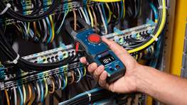 Un multimètre Bosch Professional bleu est tenu par une main, tandis qu'il est connecté à des câbles dans un tableau électrique.
