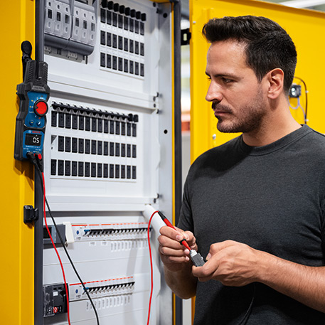 Un homme aux cheveux foncés portant un t-shirt gris travaille sur un tableau électrique. Il tient un appareil de mesure à la main tout en connectant des câbles avec un fil rouge et un fil noir. Le tableau a un extérieur jaune et contient de nombreux interrupteurs et fusibles de différentes couleurs. L'environnement semble industriel et bien éclairé, ce qui indique un atelier.