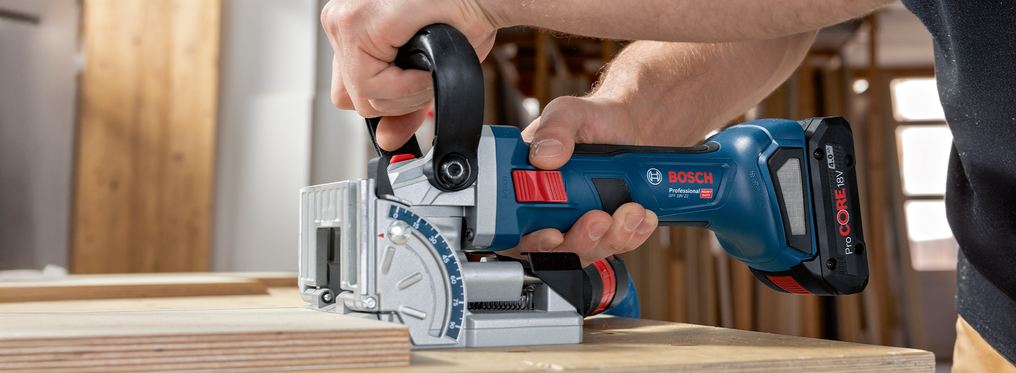 En person holder en blå, trådløs rundsav med en metalbase, der ligger på et træbræt. I baggrunden ses træplader og et lyst rum.