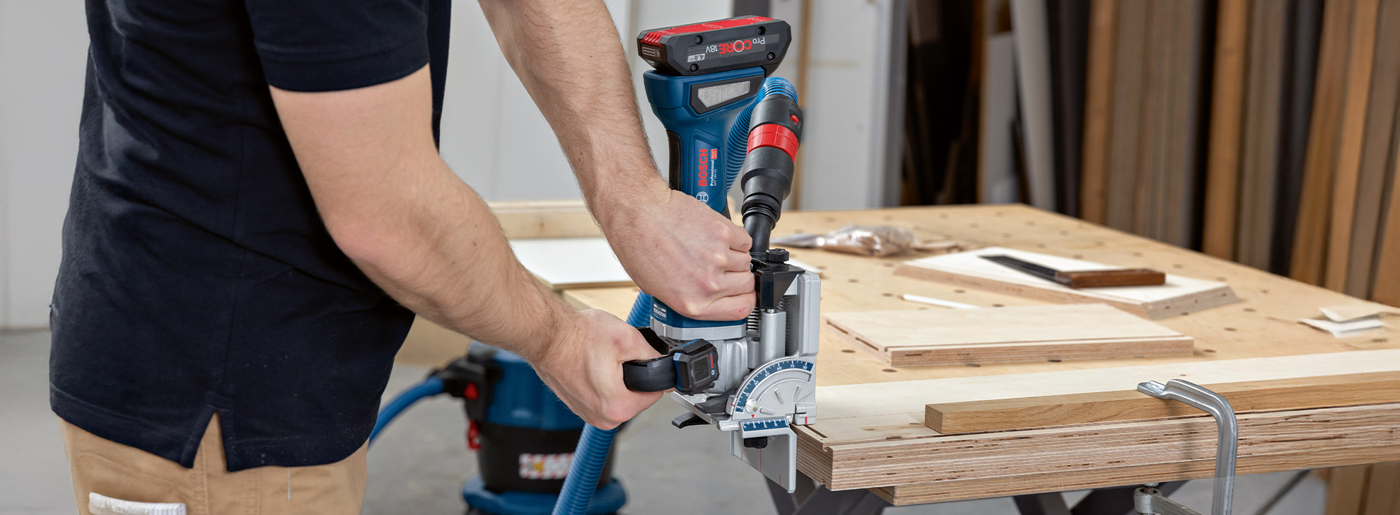 En håndværker arbejder med en blå Bosch-maskine ved et træbord, omgivet af træplader.