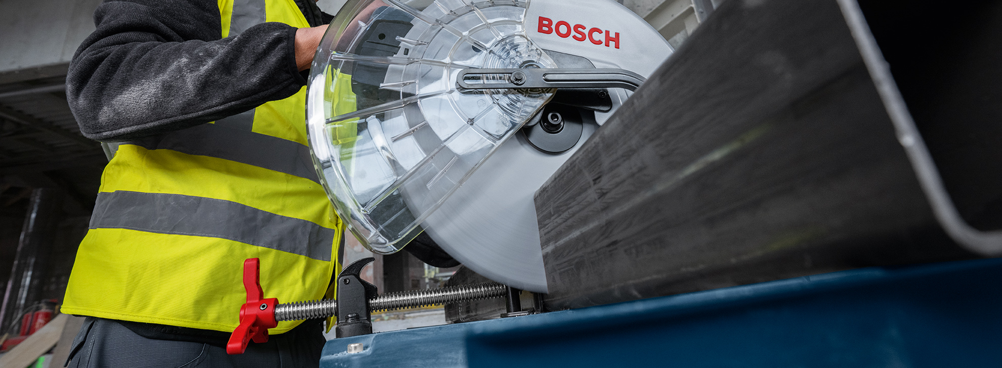 A worker in a reflective yellow vest is operating a Bosch Professional miter saw that is on a table. The saw is currently cutting a metal profile.