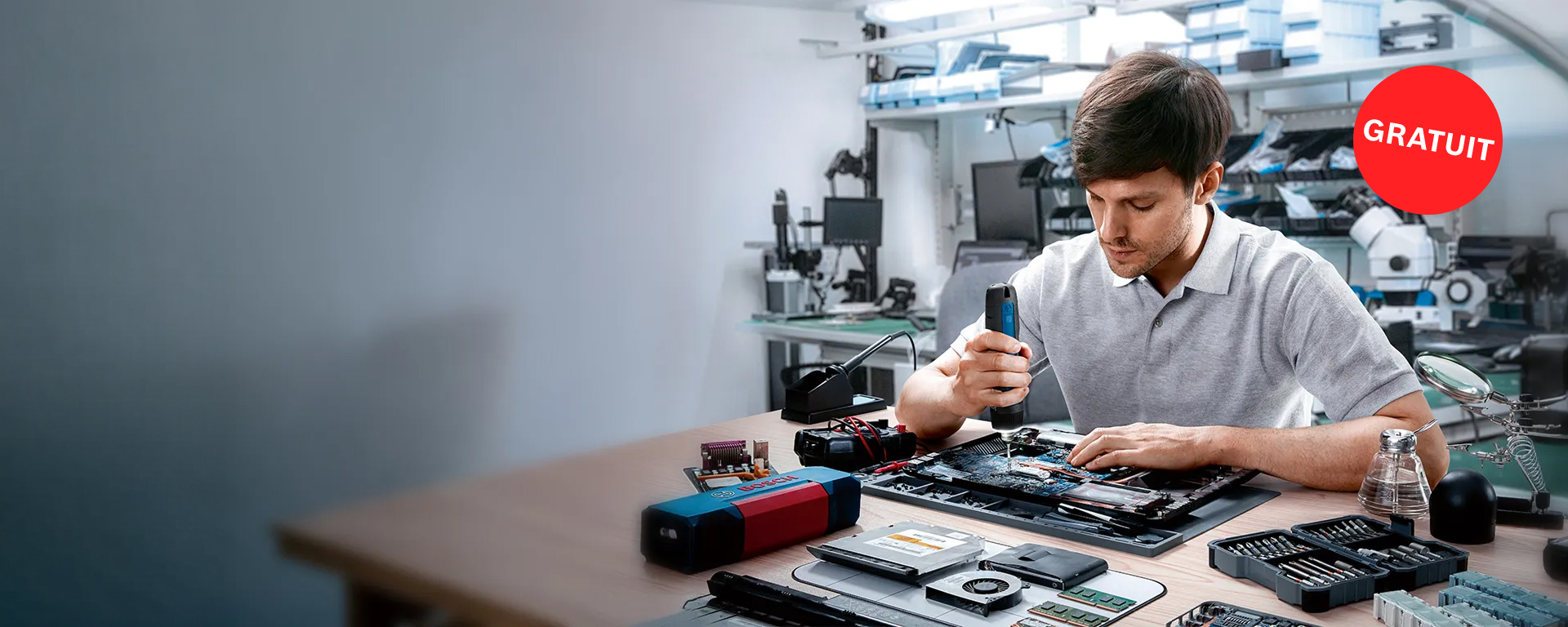 Un jeune homme en t-shirt gris répare un appareil électronique sur une table. Différents outils, dont un tournevis bleu Bosch Professional, sont éparpillés. En arrière-plan, des appareils techniques et des postes de travail sont visibles. Un symbole rouge "GRATUIT" flotte en haut à droite de l'image.