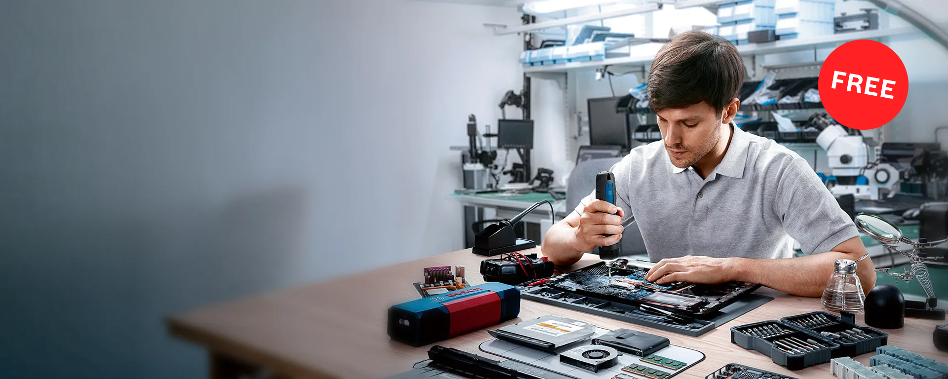 A man with short brown hair is working with a screwdriver on an open laptop. On the table are tools, including a red and blue case, as well as various components. In the background, several computers and technical devices are visible. The atmosphere is technical and focused.