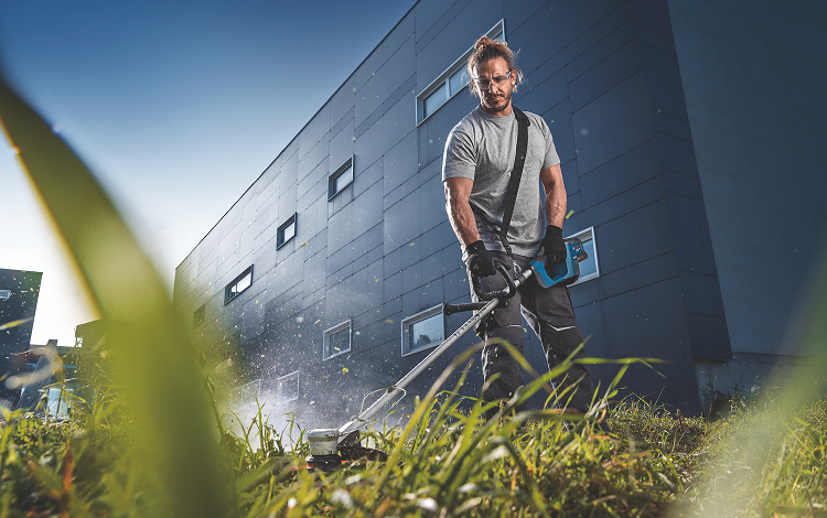 Un hombre con pantalones cortos y camiseta utiliza un cortacésped a batería Bosch Professional en un césped. Los trozos de hierba vuelan por el aire mientras trabaja concentrado. Al fondo se ve un edificio moderno y gris con ventanas. La atmósfera es activa y dinámica.