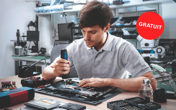 Un homme en polo gris répare une carte mère d'ordinateur portable avec un tournevis bleu Bosch Professional. Sur la table, des outils, un fer à souder et un multimètre sont visibles. En arrière-plan, d'autres appareils techniques et matériaux de travail sont visibles. L'atmosphère est concentrée et professionnelle.