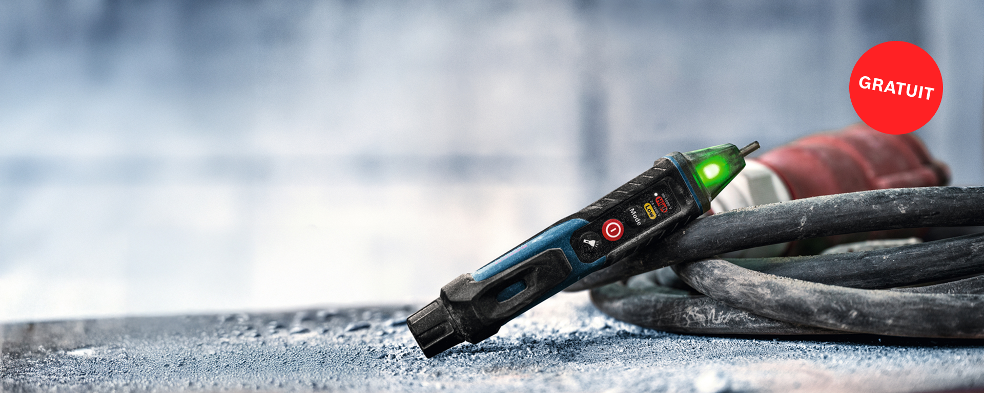 Un multimètre Bosch Professional noir et bleu est posé sur une surface grise et poussiéreuse. Une lumière verte s'allume.