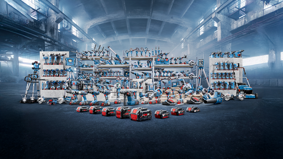 Une disposition d'outils Bosch Professional dans un grand espace industriel. Des outils bleus et gris sont répartis sur des étagères et sur le sol, avec divers appareils tels que des perceuses, des scies et des batteries. L'environnement est brut et minimaliste, avec un sol sombre et de grandes fenêtres laissant entrer une lumière tamisée.