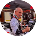 Un uomo con casco e tuta da corsa fa segno di pollice in su davanti a una tenda e attrezzi.