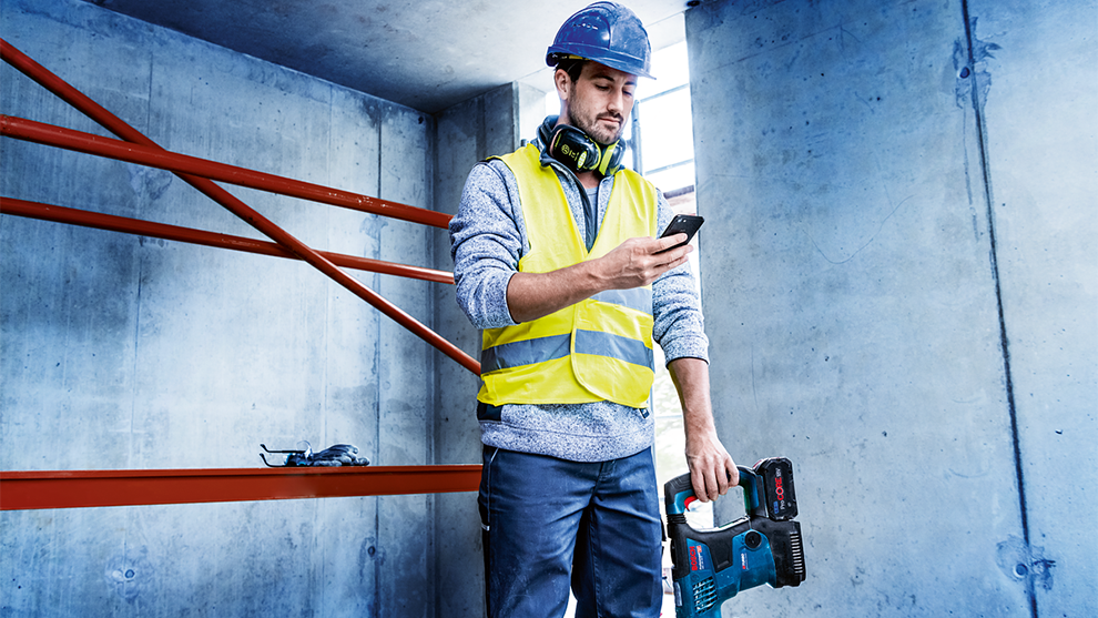 Мужчина в каске и светоотражающем жилете смотрит на свой телефон, держа инструмент Bosch.