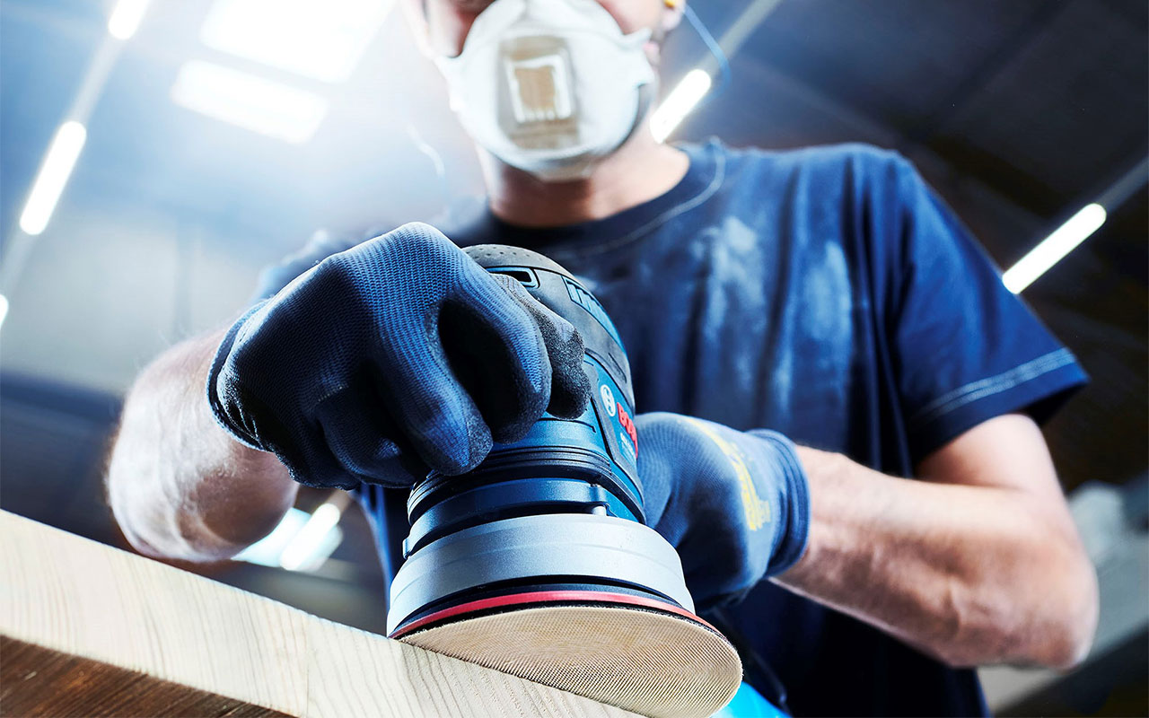 En hantverkare med blå handskar slipar trä med en Bosch-enhet.