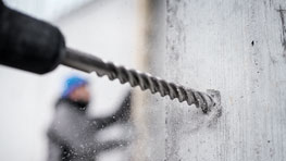 En borrmaskin genomborrar en grå vägg, i bakgrunden en person med hjälm.