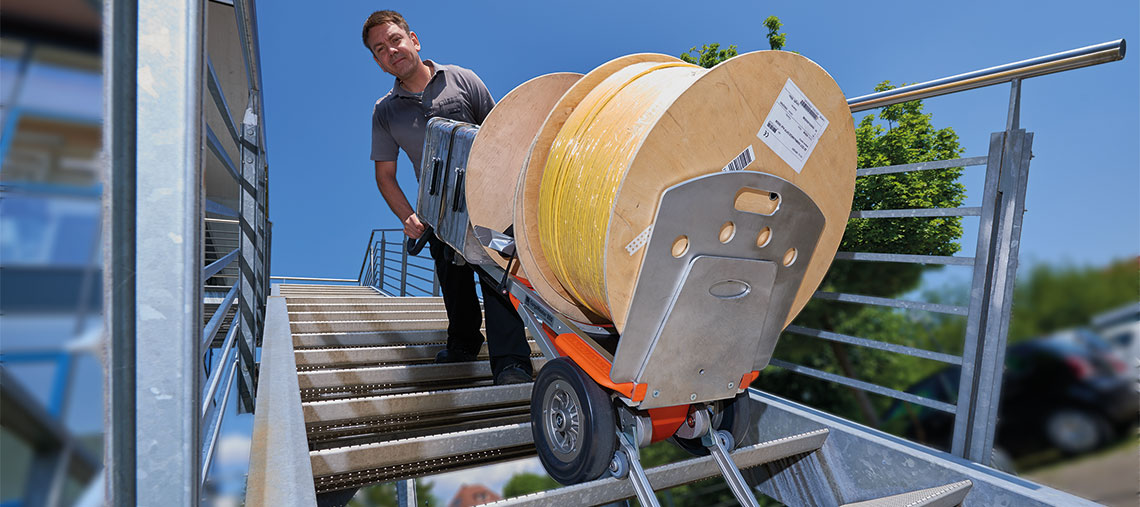 Muž táhne cívku kabelu nahoru po schodech s ručním vozíkem.