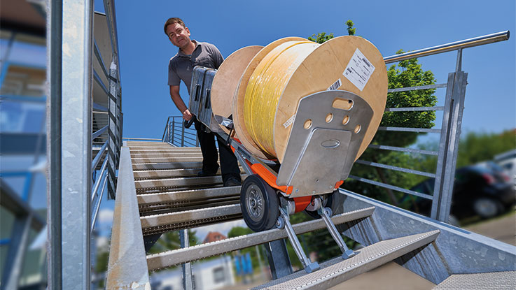 Muž táhne vozík s kabelovými cívkami dolů po schodech.