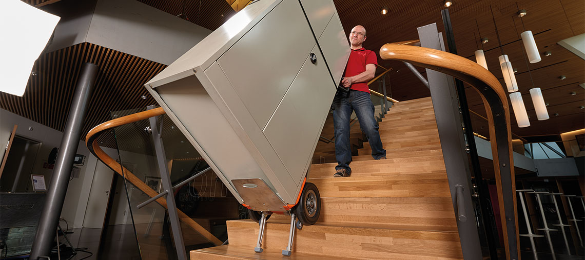 Muž nese stříbrnou skříň po dřevěném schodišti.