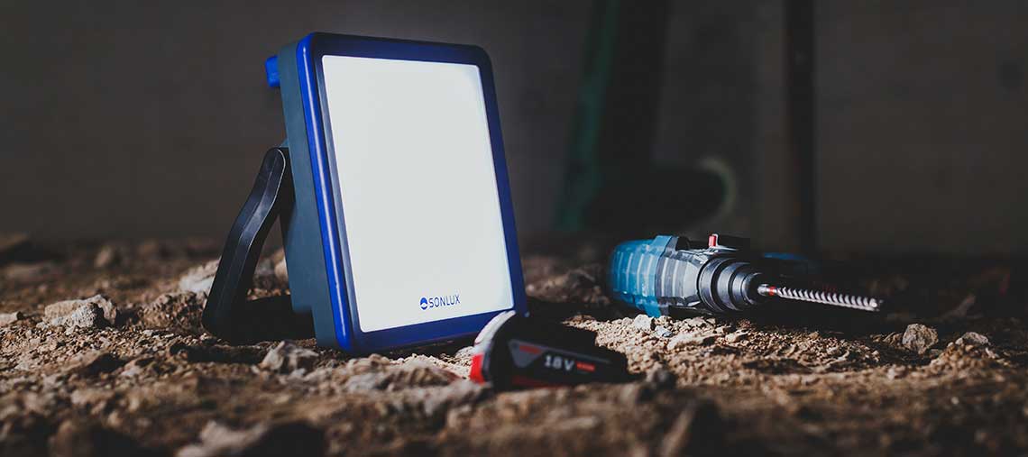 Modrá stavební lampa SONLUX stojí na podlaze vedle akumulátorové vrtačky.