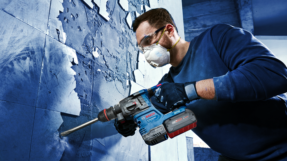 A man is using a blue Bosch hammer drill on a blue wall.