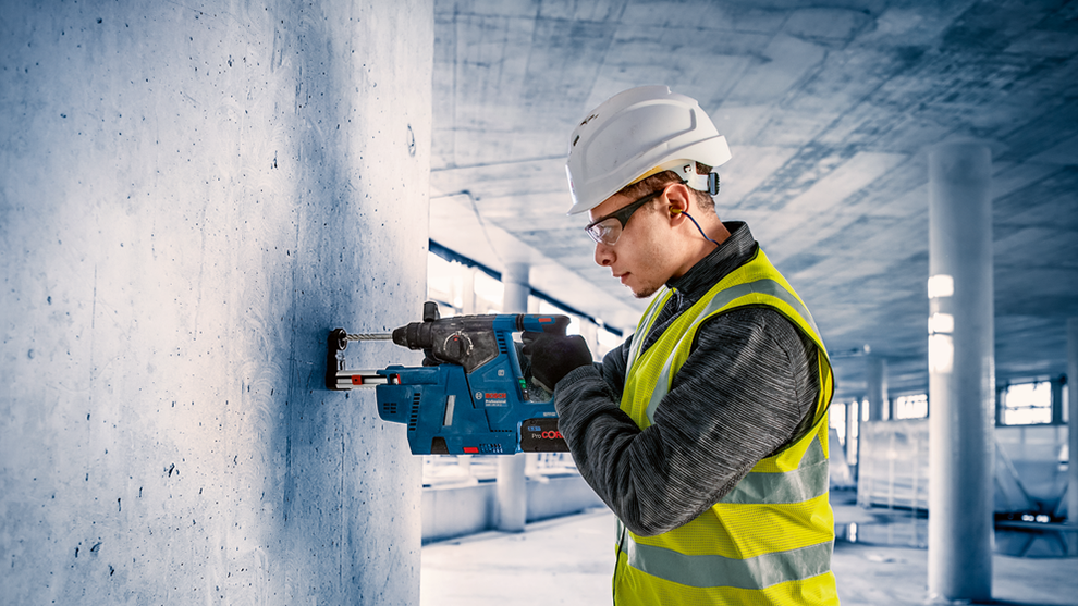 A worker wearing a helmet and vest is drilling with a blue Bosch tool.